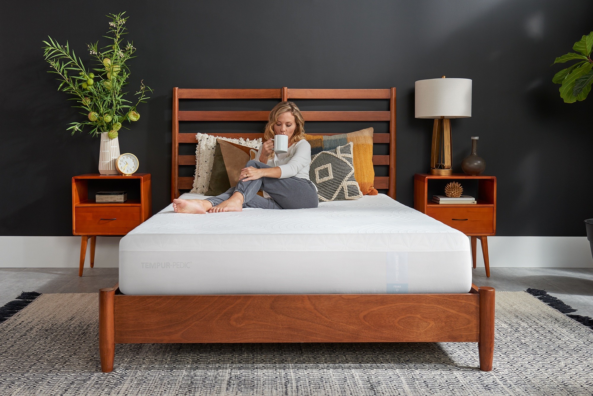 Woman sipping coffee on a mattress that is covered by a tempur-mattress protector on wooden bedroom furniture in styled bedroom