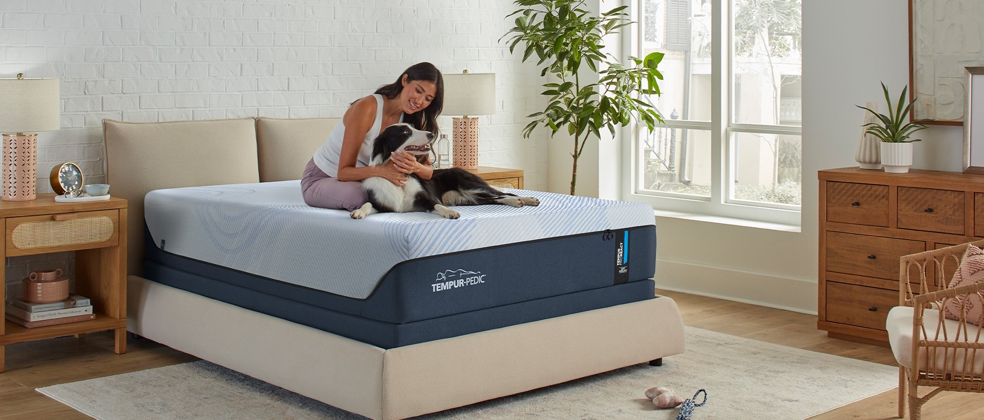 A woman smiling sitting on her Tempur React mattress while petting a long-haired black and white dog